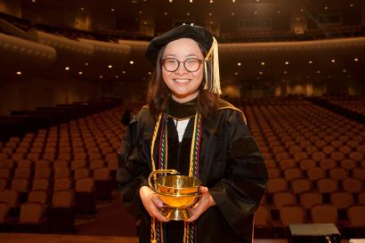 La standing in cap and gown and holding golden bowl