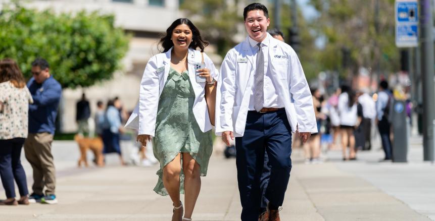 two students wearing white coats walk down a sidewalk.