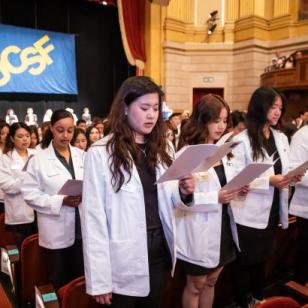 large group of students wearing white coats facing the audience in a theater holding paper