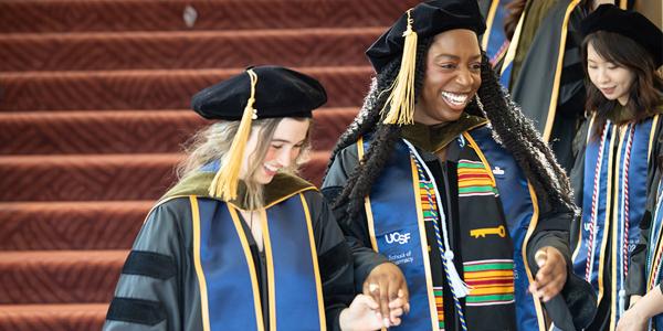 two smiling graduates in cap and gown hold hands while descending stairs
