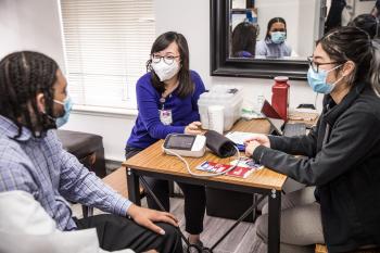 Chong and Zhou speaking with Hogan while seated at a table with a blood pressure reader
