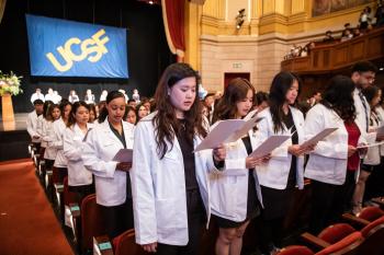large group of students wearing white coats facing the audience in a theater holding paper