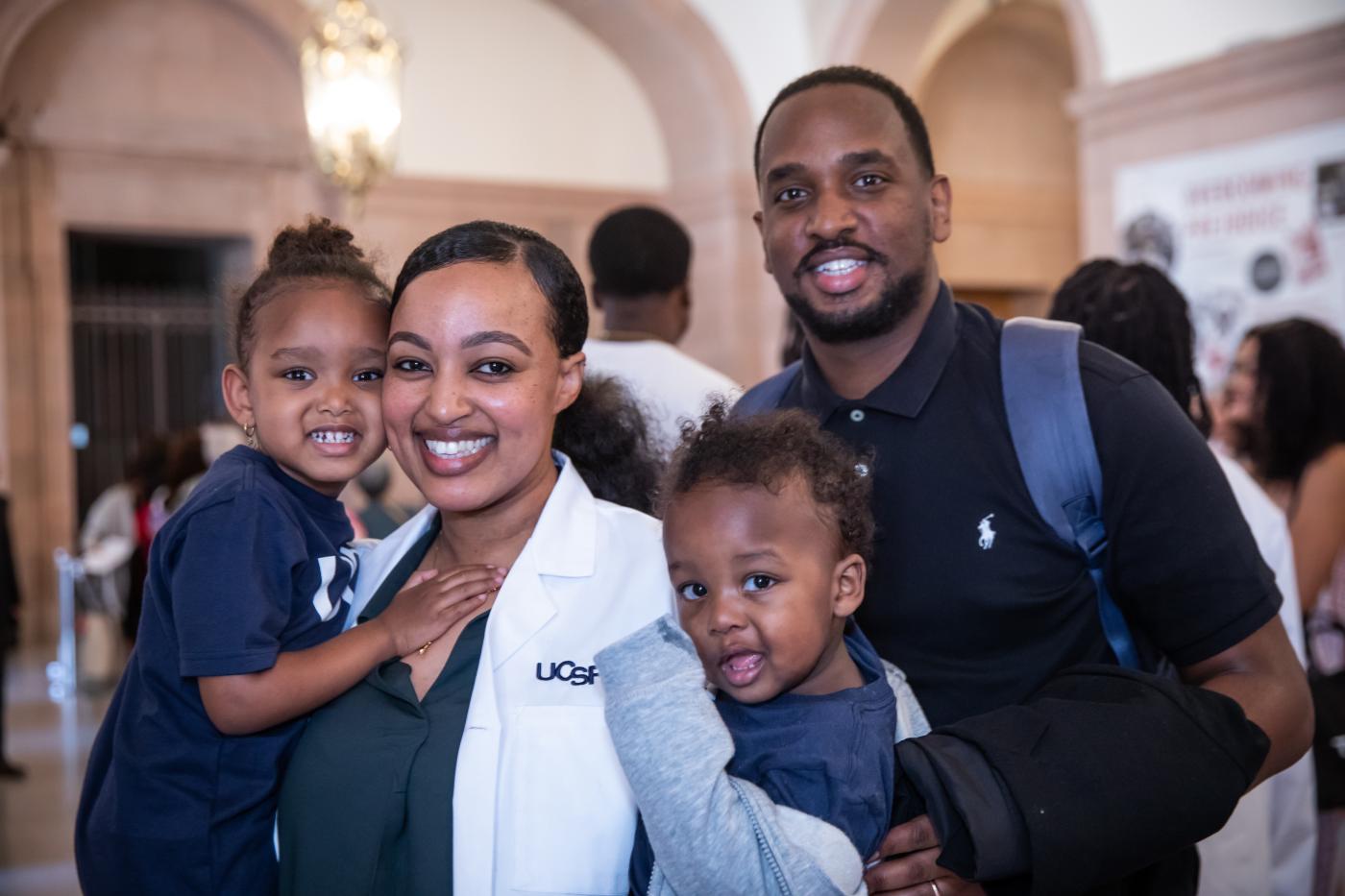 Woman in white coat holds two children braced by a man behind.